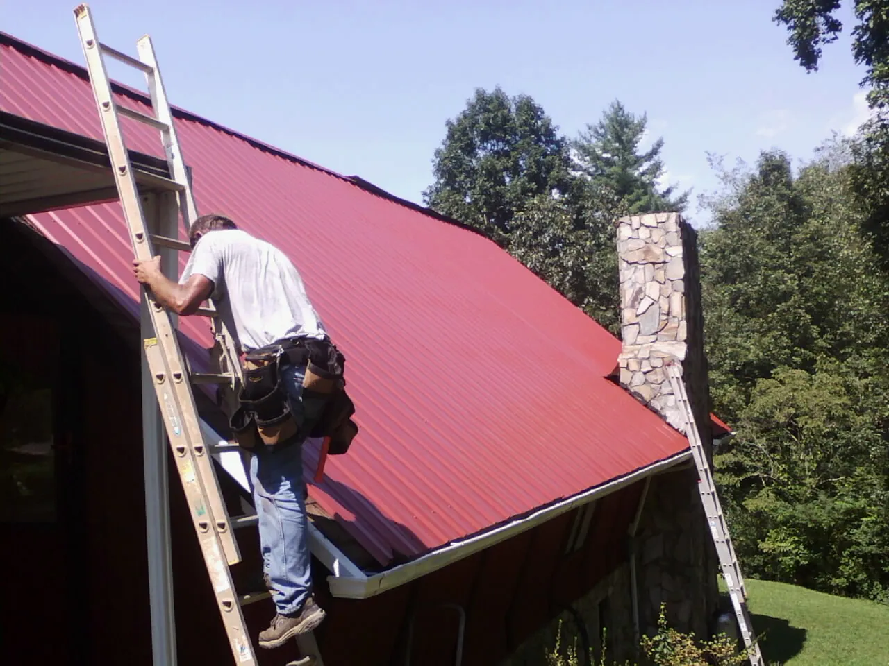 Roofer on a ladder beside a red metal roof and stone chimney — Soffit & Fascia Repair by Roofing Brothers in Towson